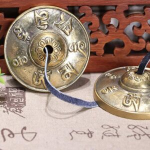 Close-up side view of brass tingsha bells showing engraved floral pattern and sacred mantras with Tibetan script backdrop