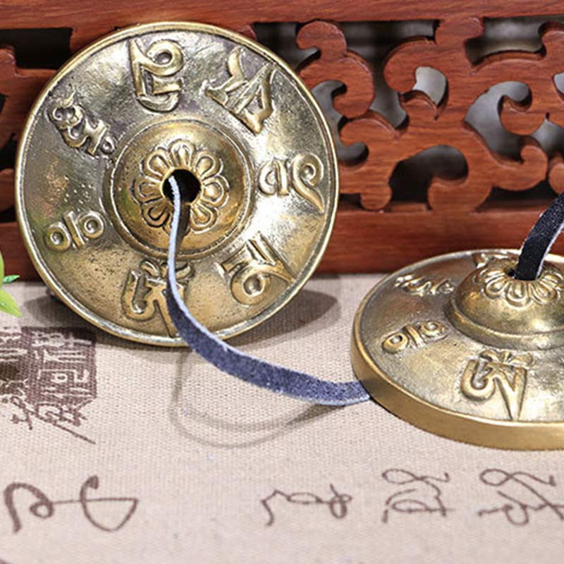 Close-up side view of brass tingsha bells showing engraved floral pattern and sacred mantras with Tibetan script backdrop