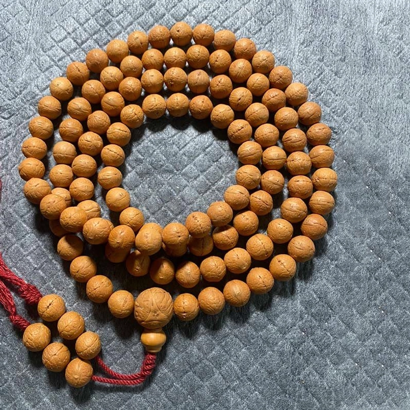 108 Bodhi seed mala beads bracelet with light tan round beads and red fabric tassel pendant displayed on quilted surface