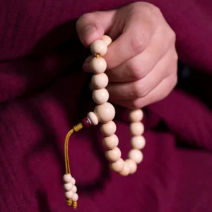 Abelia Biflora wood wrist mala with red accent beads held in hand, showcasing cream-colored natural wood beads and yellow tassel on dark red fabric background