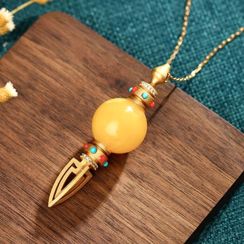 24K gold plated amber dorje vajra pendant with red-green gemstone inlay, top view on wooden coaster with teal fabric