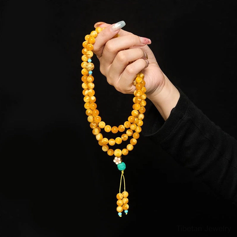 Handheld amber mala prayer beads worn in hand with yellow cord and turquoise tassel charm against dark background