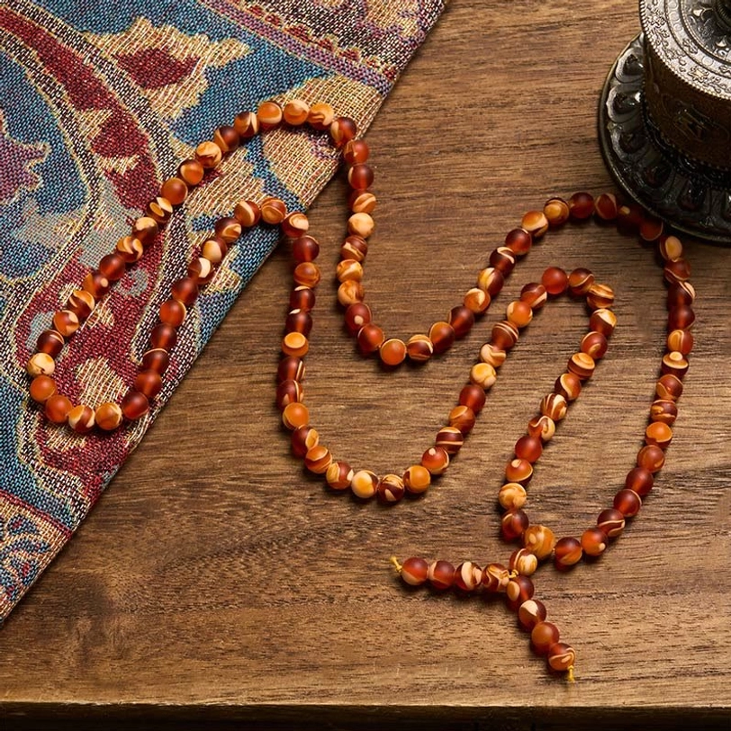 Long strand amber prayer mala draped on wooden table with patterned fabric, showcasing full length and rustic aesthetic