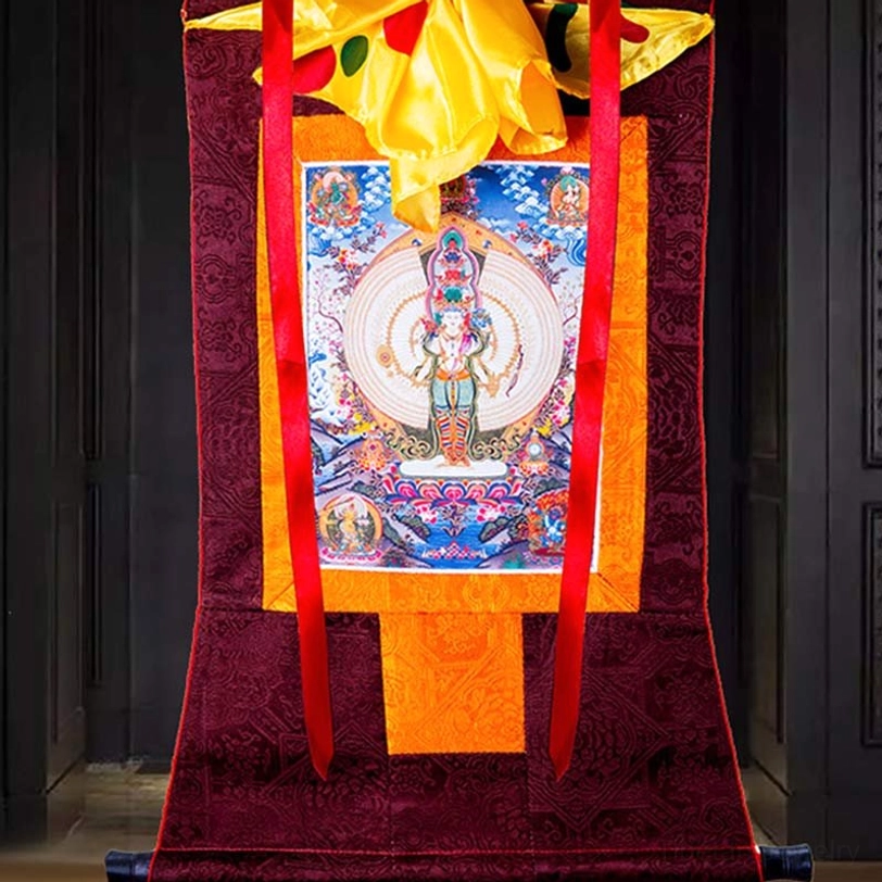 Tibetan Buddhist thangka scroll art with dark red brocade trim and yellow top fabric displayed against dark paneled interior wall
