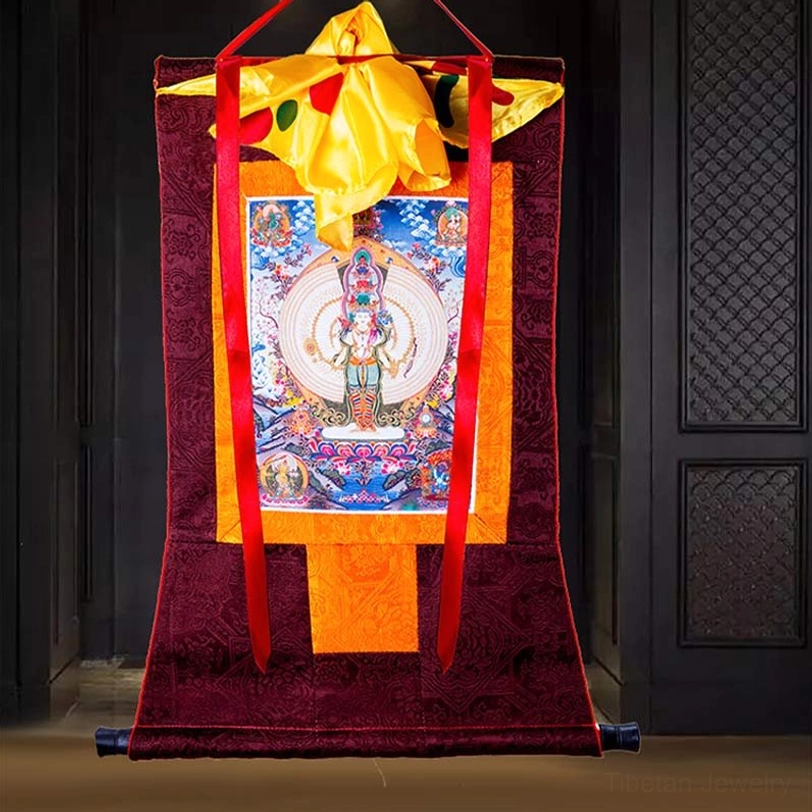 Thousand-armed Avalokitesvara thangka framed scroll with dark red woven fabric border and red hanging straps on wooden panel background