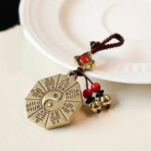 Feng Shui Bagua brass keychain with engraved eight trigrams and Yin Yang center symbol, displayed on white ceramic plate with decorative bead tassels