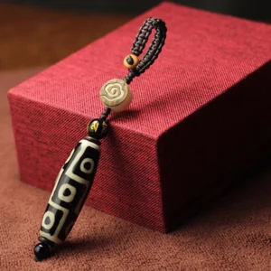 Single black Nine-Eye Dzi Bead pendant with distinctive white concentric eye pattern, braided rope attachment, presented on red gift box and brown fabric backdrop
