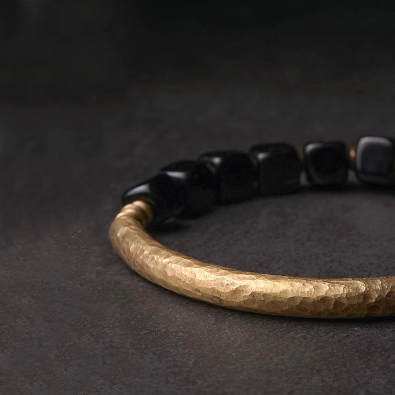 Full black obsidian couple bracelet side lay on gray background, hammered brass band with cube stone beads visible