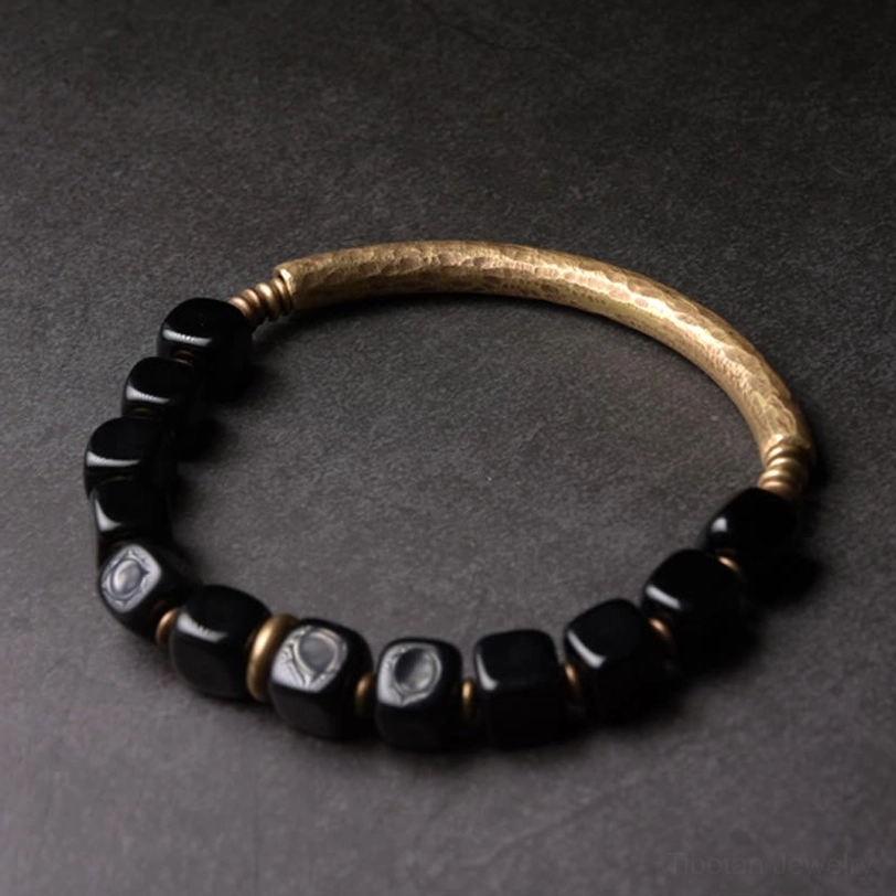 Black obsidian bracelet overhead flat lay with engraved logo beads, polished stones and hammered brass band on gray background