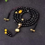 108 black obsidian mala bracelet with lotus tassel, flat lay display showing glossy round beads with gold hardware and yellow accent beads