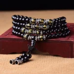 Black Obsidian 108-bead mala bracelet with gold-carved mantra beads and silver tassel on wooden display stand, top-down spiritual jewelry photography