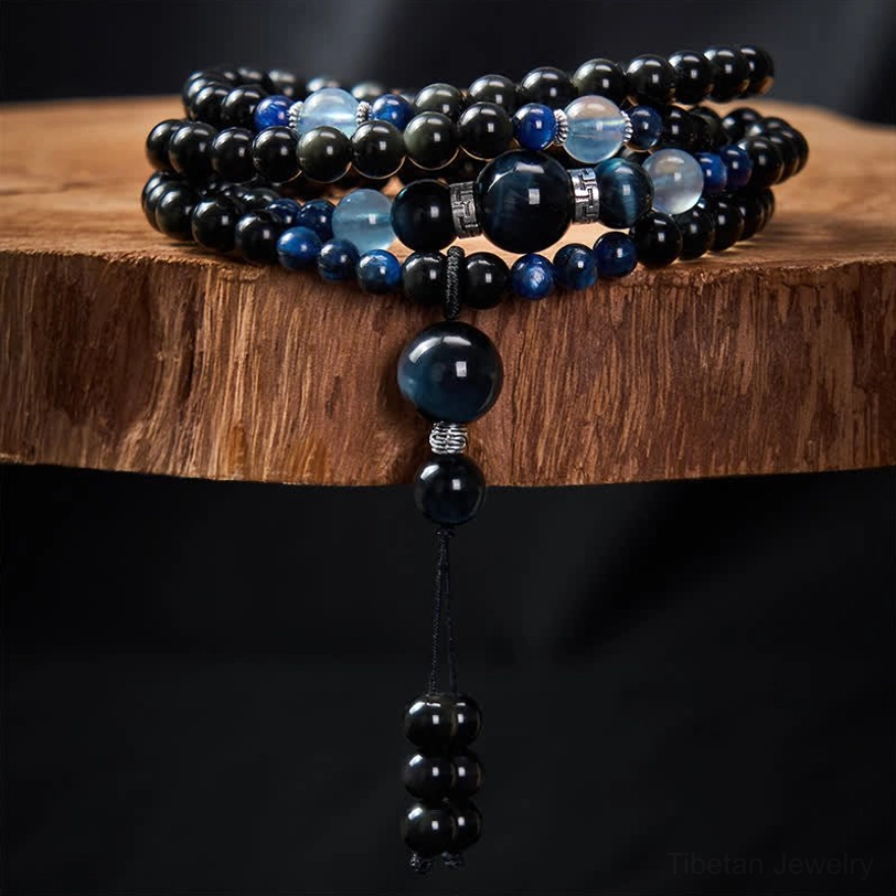 Coiled Black Obsidian 108 bead mala with blue accent beads and decorative tassel, stacked on wooden display slab close-up