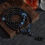 Black Obsidian 108 bead mala necklace flat lay on wooden display board, showcasing complete strand with blue accent beads and tassel pendant from above