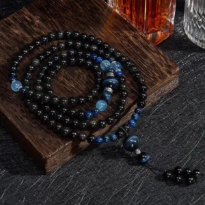 Black Obsidian 108 bead mala necklace flat lay on wooden display board, showcasing complete strand with blue accent beads and tassel pendant from above