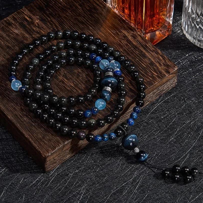Black Obsidian 108 bead mala necklace flat lay on wooden display board, showcasing complete strand with blue accent beads and tassel pendant from above