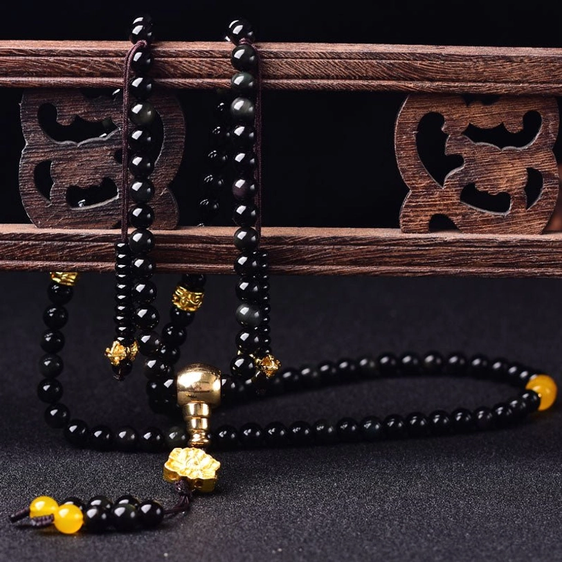 Black obsidian prayer mala hanging vertically from carved wooden shelf, displaying full length with gold fittings and lotus tassel