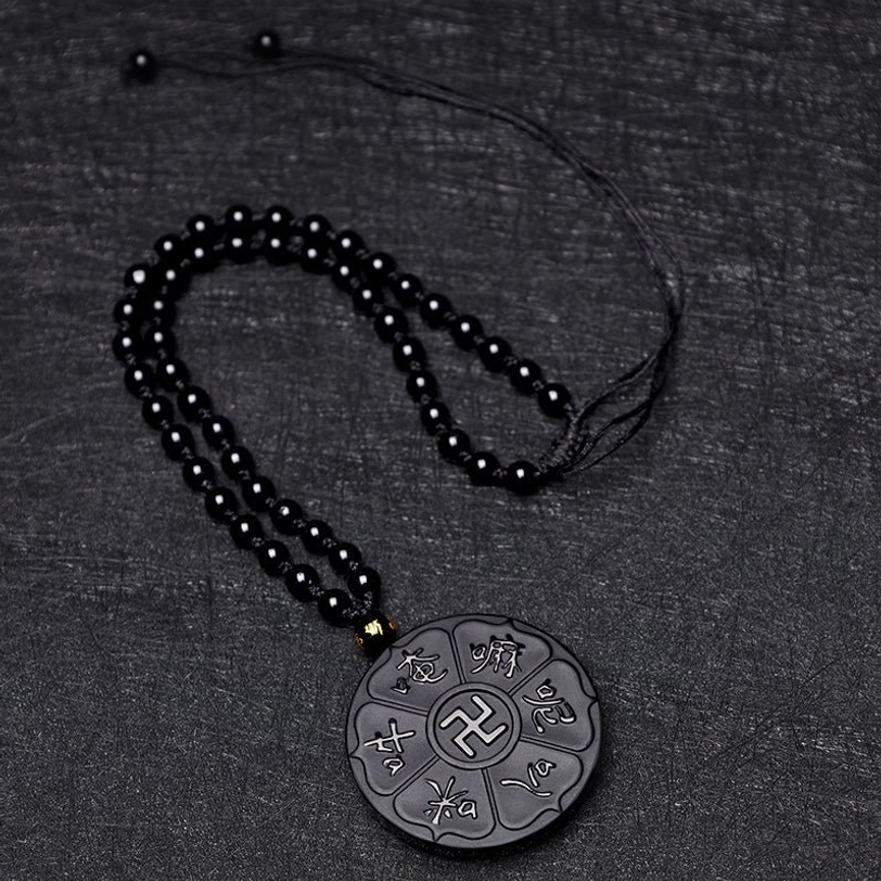 Black obsidian bead necklace with handcrafted pendant featuring silver-engraved Tibetan mantra and swastika, flat-lay dark fabric background