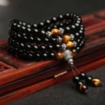 Black obsidian tiger eye mala bracelet 108 beads flat lay with silver accents and tassel, displayed on red wooden tray