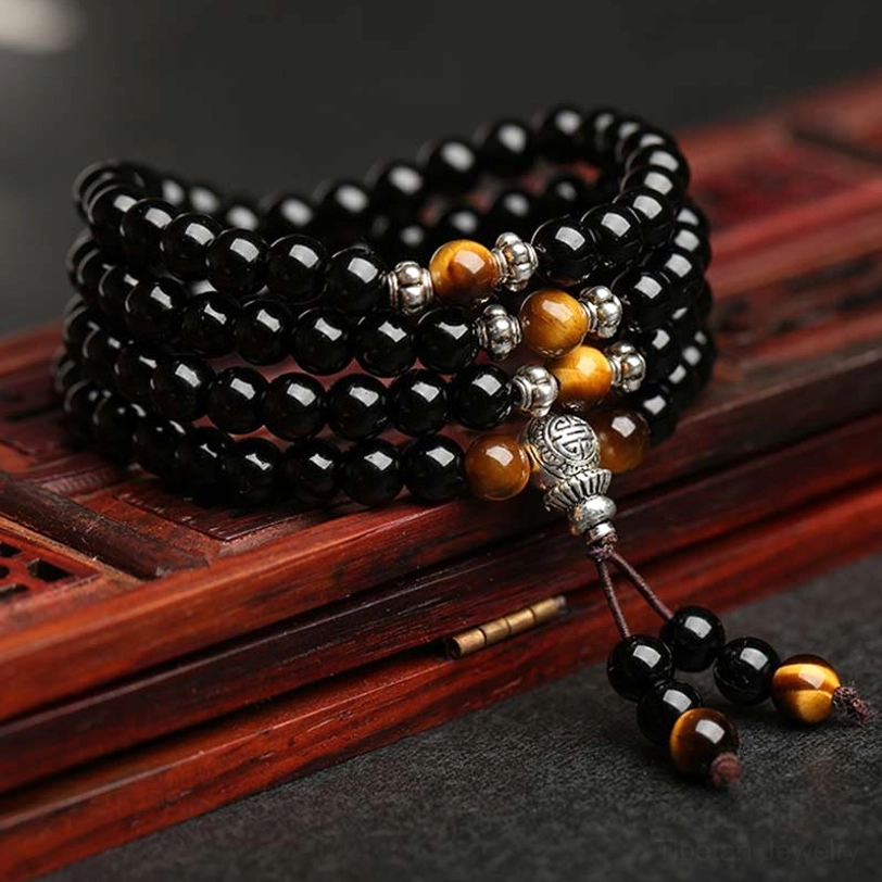 Black obsidian tiger eye mala bracelet 108 beads flat lay with silver accents and tassel, displayed on red wooden tray