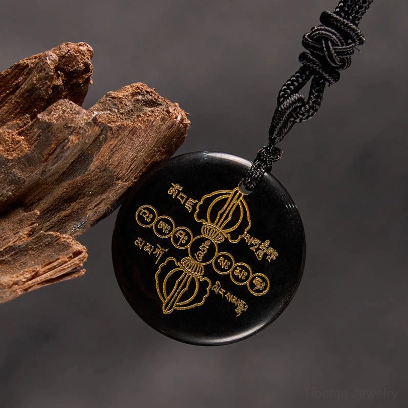 Side angle view of black obsidian Vajra Dorje necklace pendant leaning against weathered wood, showing golden religious engravings