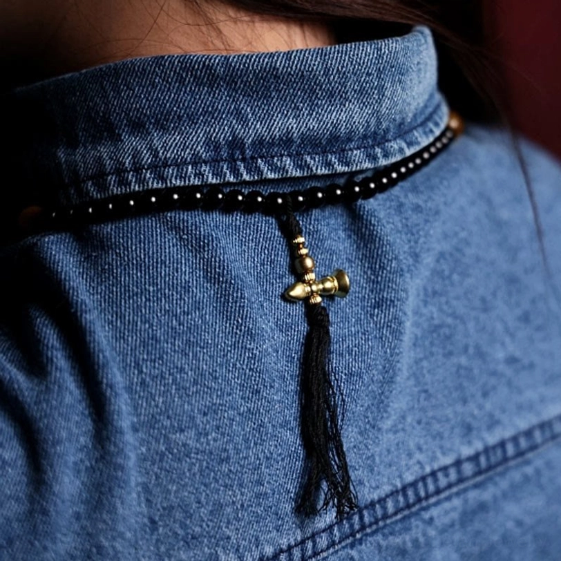 Wearable black onyx 108 mala bracelet with vintage brass accents and black cotton tassel bell pendant worn on denim shirt collar