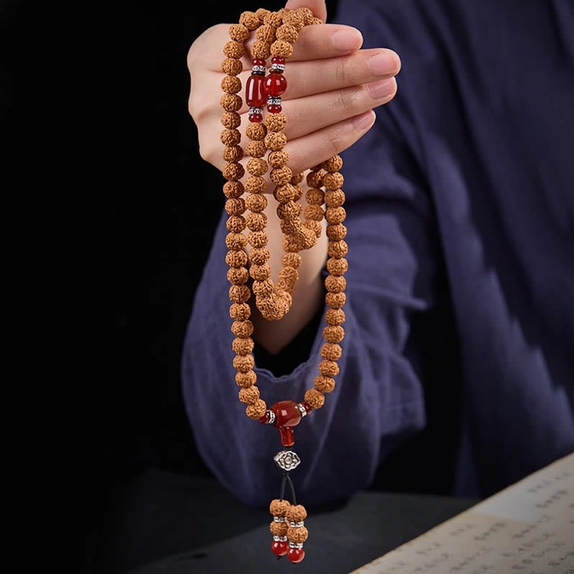 Side close-up of held bodhi seed mala with tassel pendant, displaying brown seeds with red and silver accents in hand