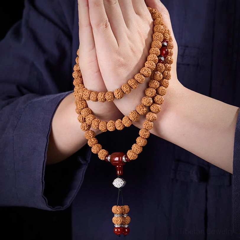 Front view of bodhi seed mala wrapped between two hands showing detailed tassel pendant, brown seeds with red agate and silver components