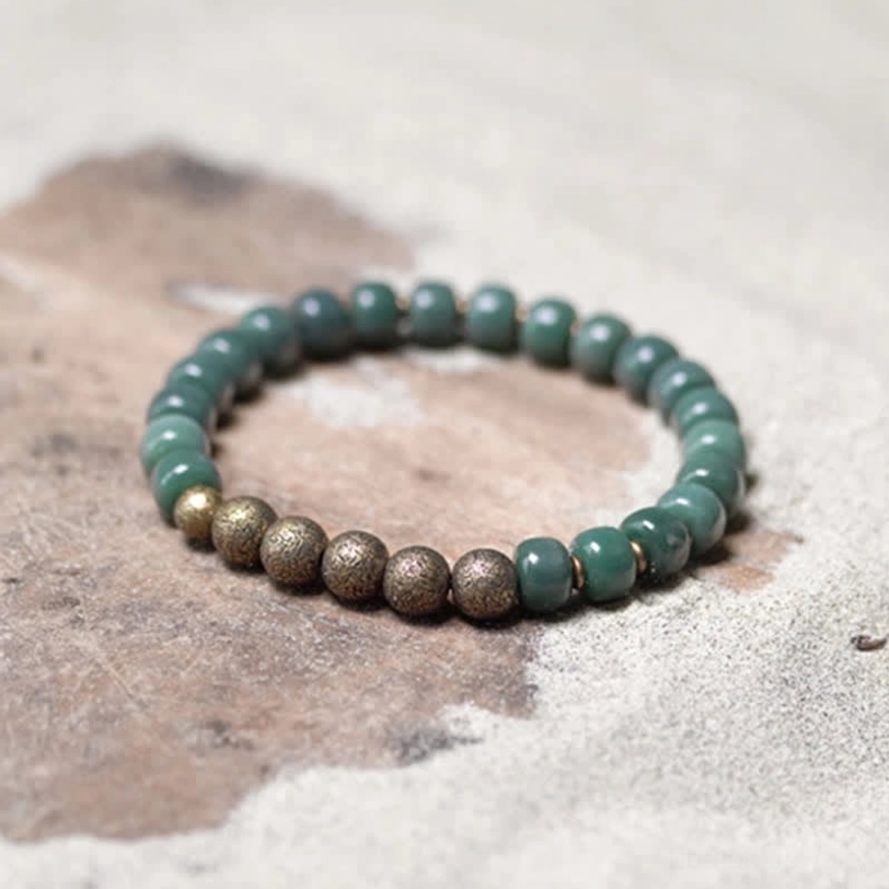 Overhead flat lay of bodhi seed copper bracelet on sand-stained wooden background, displaying matte dark green beads with bronze spacer beads
