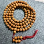 108 Bodhi seed mala beads bracelet with red tassel displayed on grey quilted fabric, light brown natural seeds