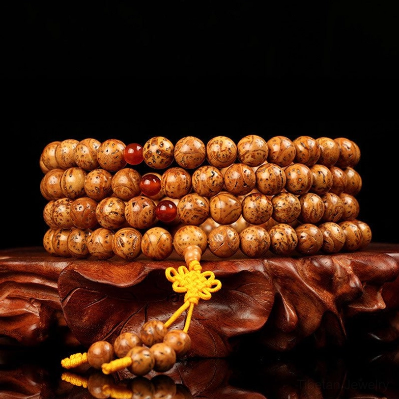 Tibetan bodhi seed mala bracelet with 108 beads displayed on carved wooden stand, showing natural spotted brown seeds with red agate spacers and yellow silk tassel