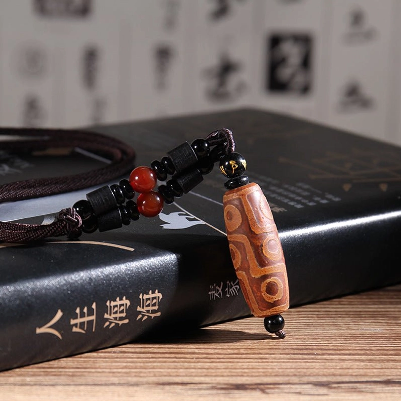 Brown nine-eye dzi bead pendant closeup showing etched pattern with black braided cord and red agate