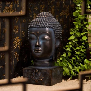 Bronze Buddha head sculpture with dark patina showing front facing view on tabletop with calligraphy backdrop and green plants