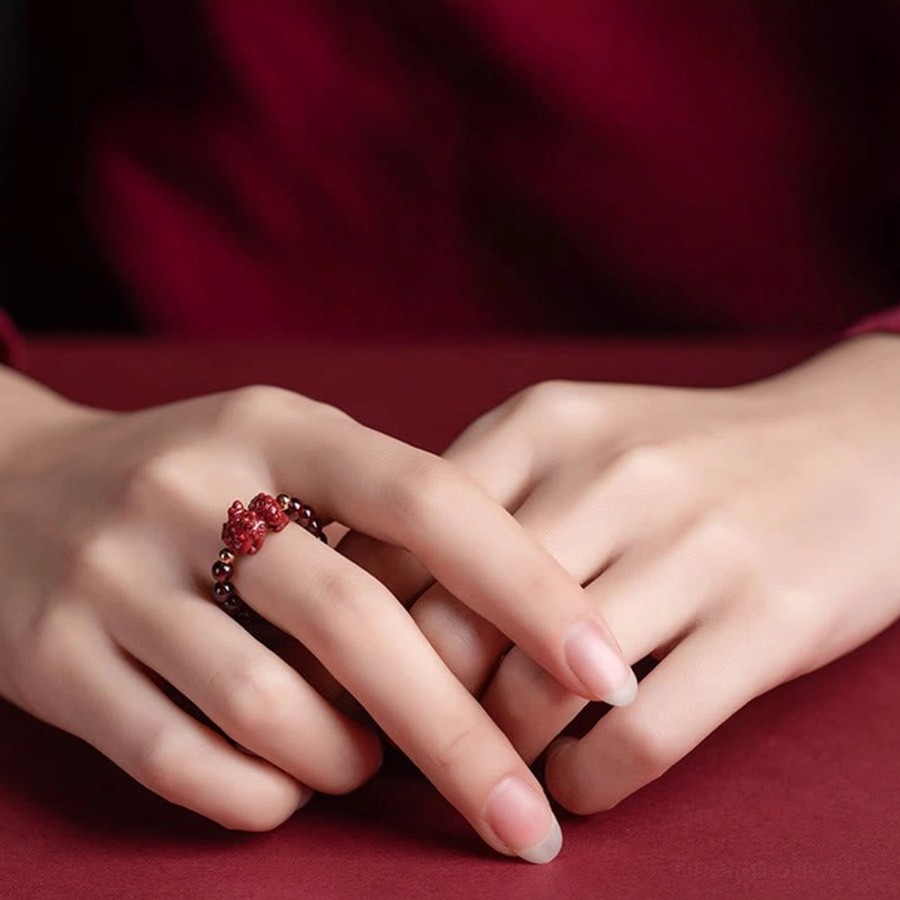 Carved cinnabar PiXiu ring with ornate gold metal spacers and detailed protective charm, eye-level view on wine red fabric