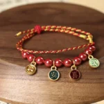 Cinnabar red string blessing bracelet with gold-tone charms displayed on wooden tray, showing intricate braided weave and natural beads