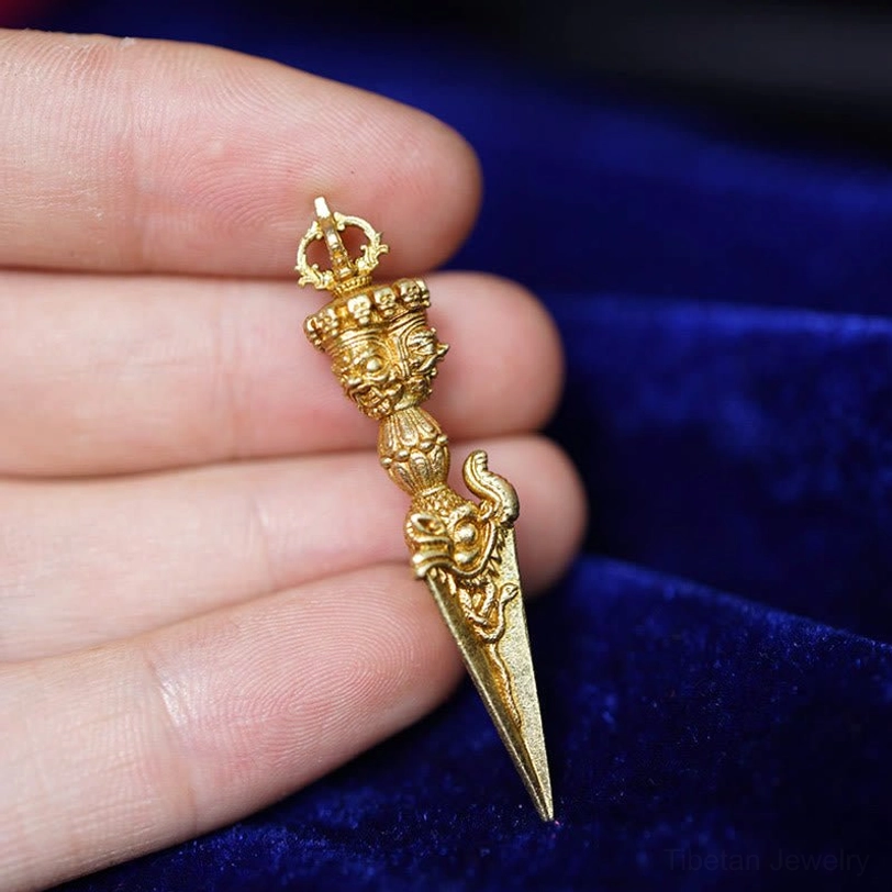 Dorje Vajra spiritual pendant in bright gold finish held in fingers against blue velvet background, ornate religious carving motifs