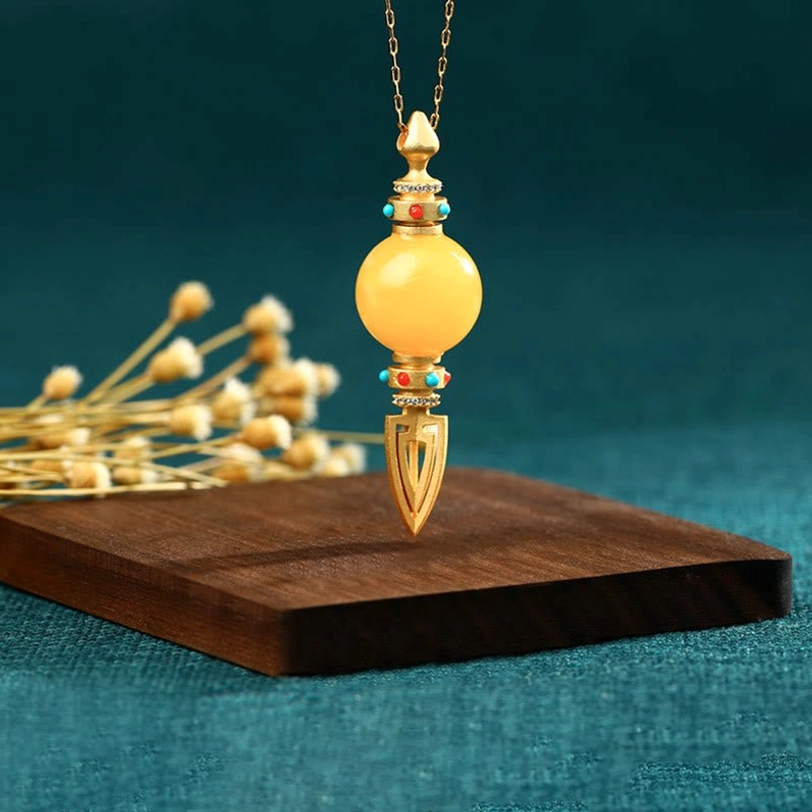 Side hanging view of 24K gold plated amber dorje vajra pendant on chain with teal background and botanical decor