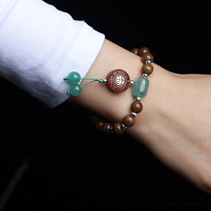 Double-strand tibetan dragon wood bracelet worn on wrist with green jade pieces, fu charm, and tassel accents on dark backdrop
