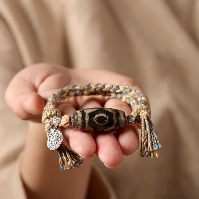 First-person close-up of Dzi bead bracelet held in palm showing full view of sacred bead, braided rope, and metal charm details