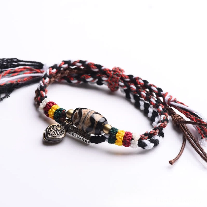 Double-wrap braided Tibetan Dzi bead bracelet with red-black woven rope, metal pendants, and loose tassel ends on brown background