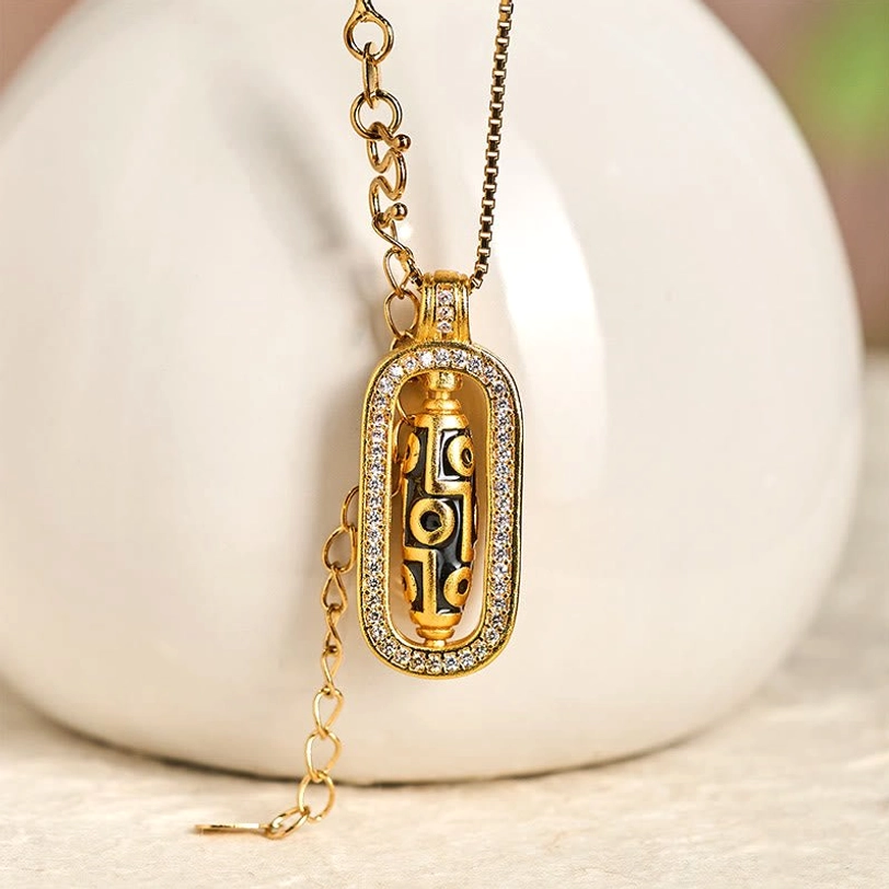 Rotatable Tibetan nine-eye dzi bead necklace with gold chain displayed beside white ceramic vase, three-quarter view