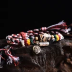 Tibetan Dzi Bead Om Mani Padme Hum braided rope bracelet with red-black-white woven cord and copper metal pendants on dark wooden display