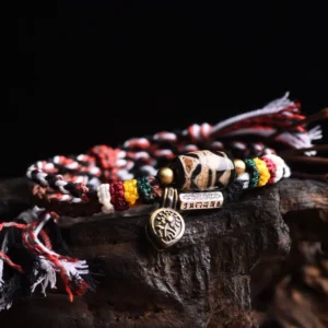 Tibetan Dzi Bead Om Mani Padme Hum braided rope bracelet with red-black-white woven cord and copper metal pendants on dark wooden display