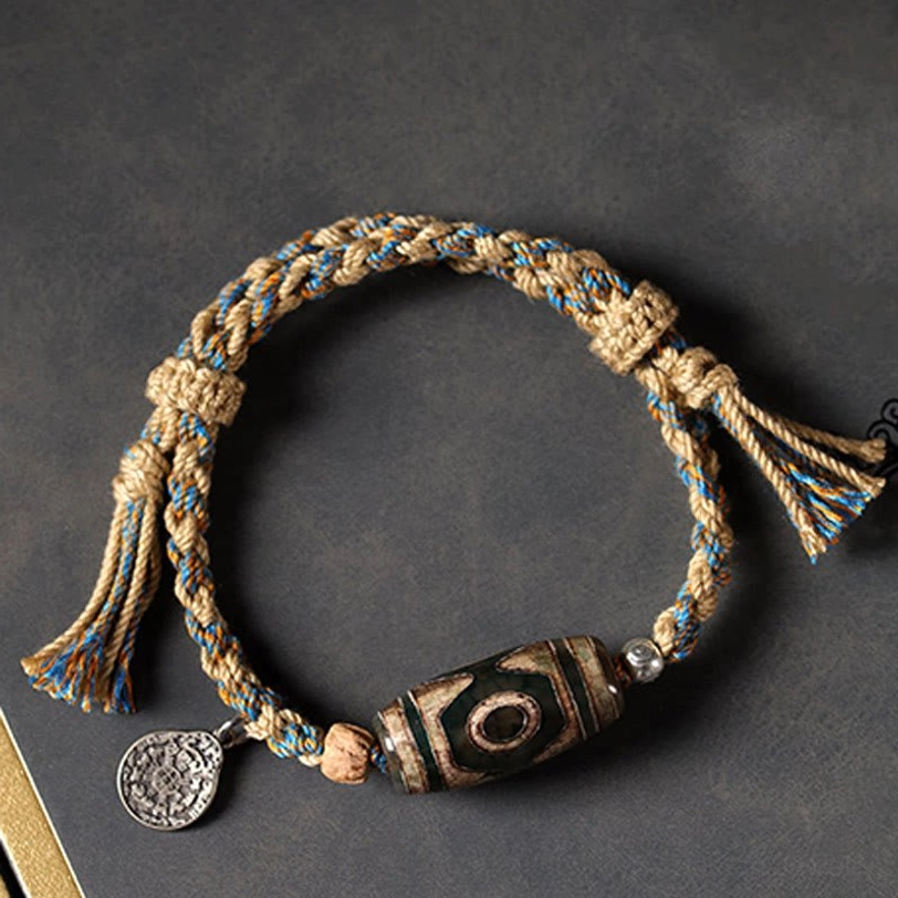 Close-up flat lay of handmade Tibetan Dzi bead bracelet showing textured woven rope, prominent protective bead, metal tag, and handcrafted knots on dark gray background