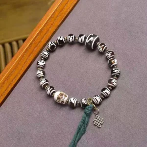 Tibetan Dzi bead and skull bracelet top view on purple fabric with wooden border, featuring green tassel and metal charm