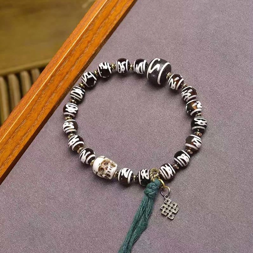 Tibetan Dzi bead and skull bracelet top view on purple fabric with wooden border, featuring green tassel and metal charm