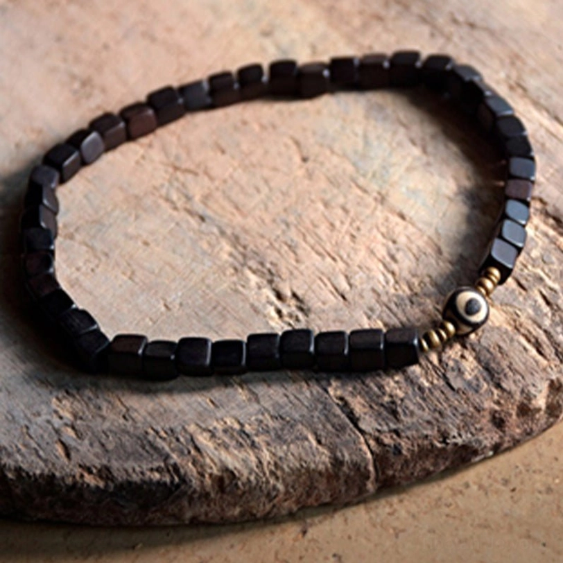 Minimalist ebony wood dzi bead couple bracelet featuring polished black square beads with gold-toned metal details and central eye bead on rough wooden surface
