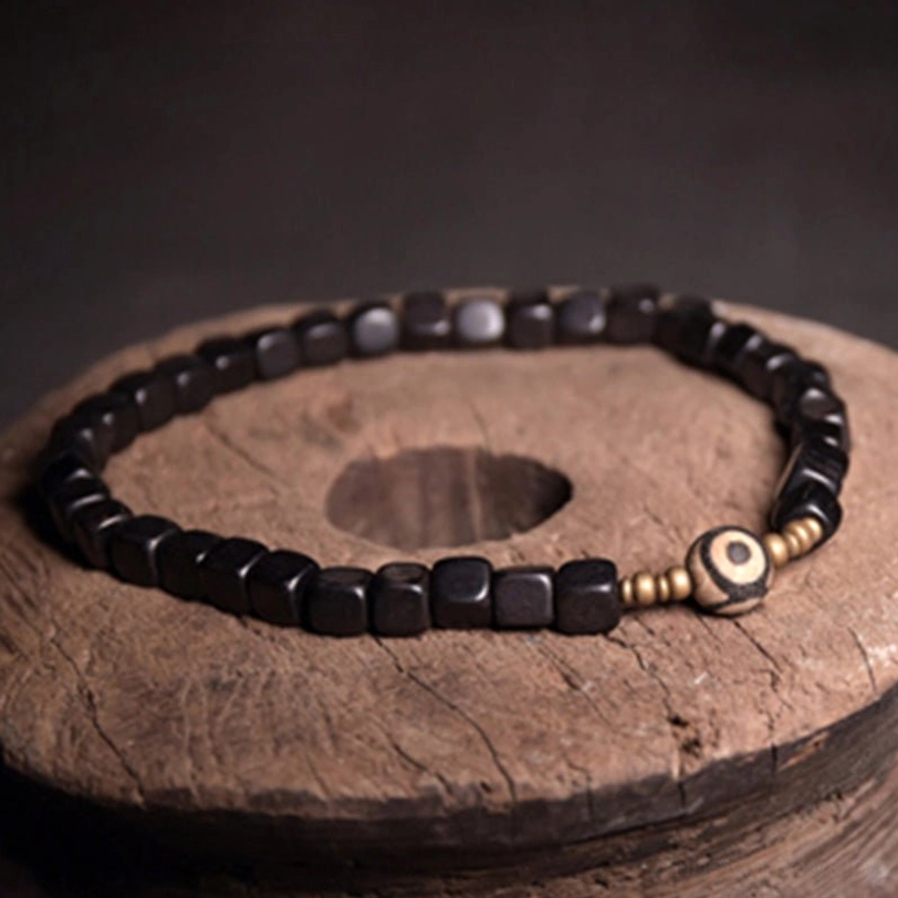 Single-wrap ebony wood dzi bead bracelet top-down flat lay showing uniform black cube beads with copper accents and central eye charm on wooden coaster