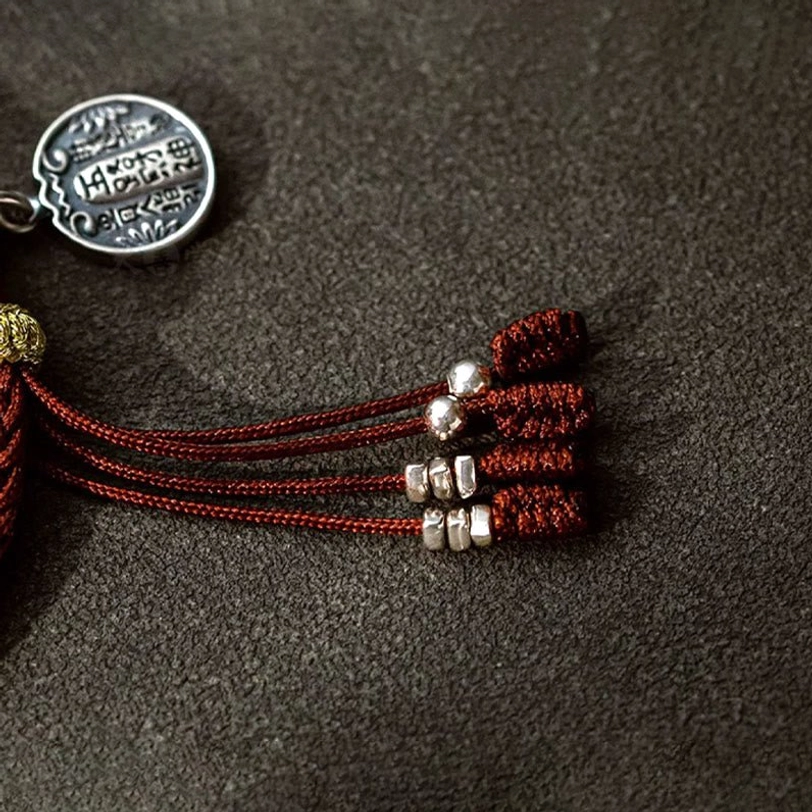 Sterling silver wealth charm with red tassel and silver bead accents, showing engraved text detail, flat lay on dark background