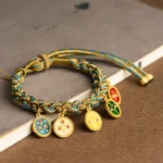 Tibetan Five God of Wealth braided string bracelet with blue-gold twisted rope and enamel deity pendants, displayed on wooden desktop with book