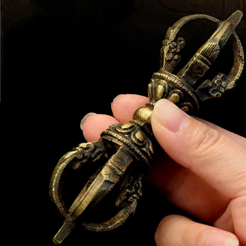 Five-prong Tibetan Vajra Dorje held by fingers against dark background, revealing detailed carved ends and aged bronze metal texture